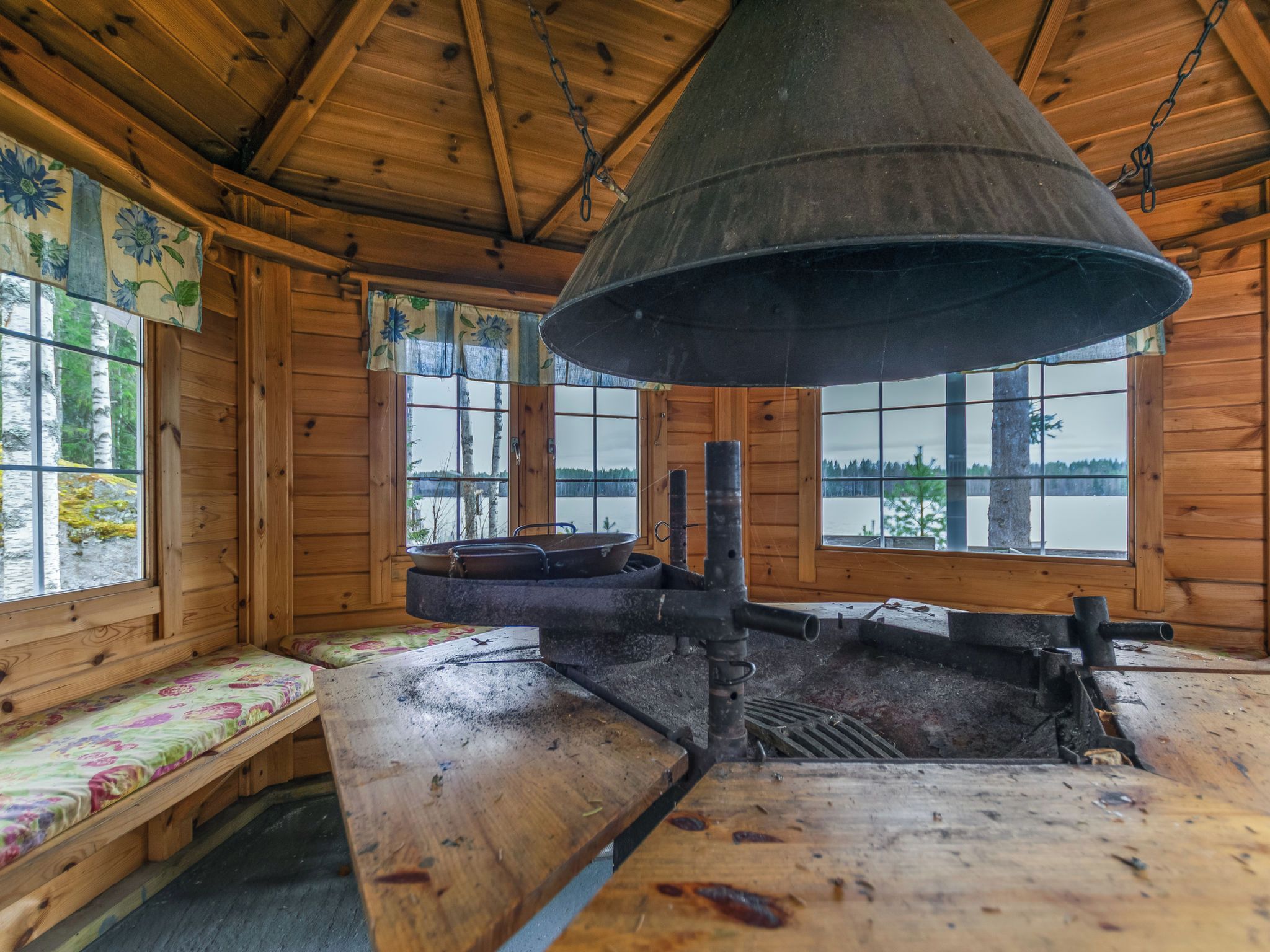 A rustic cabin interior with a large metal stove, wooden benches, and large windows offering views of a forested landscape.