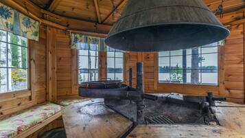 A rustic cabin interior with a large metal stove, wooden benches, and large windows offering views of a forested landscape.