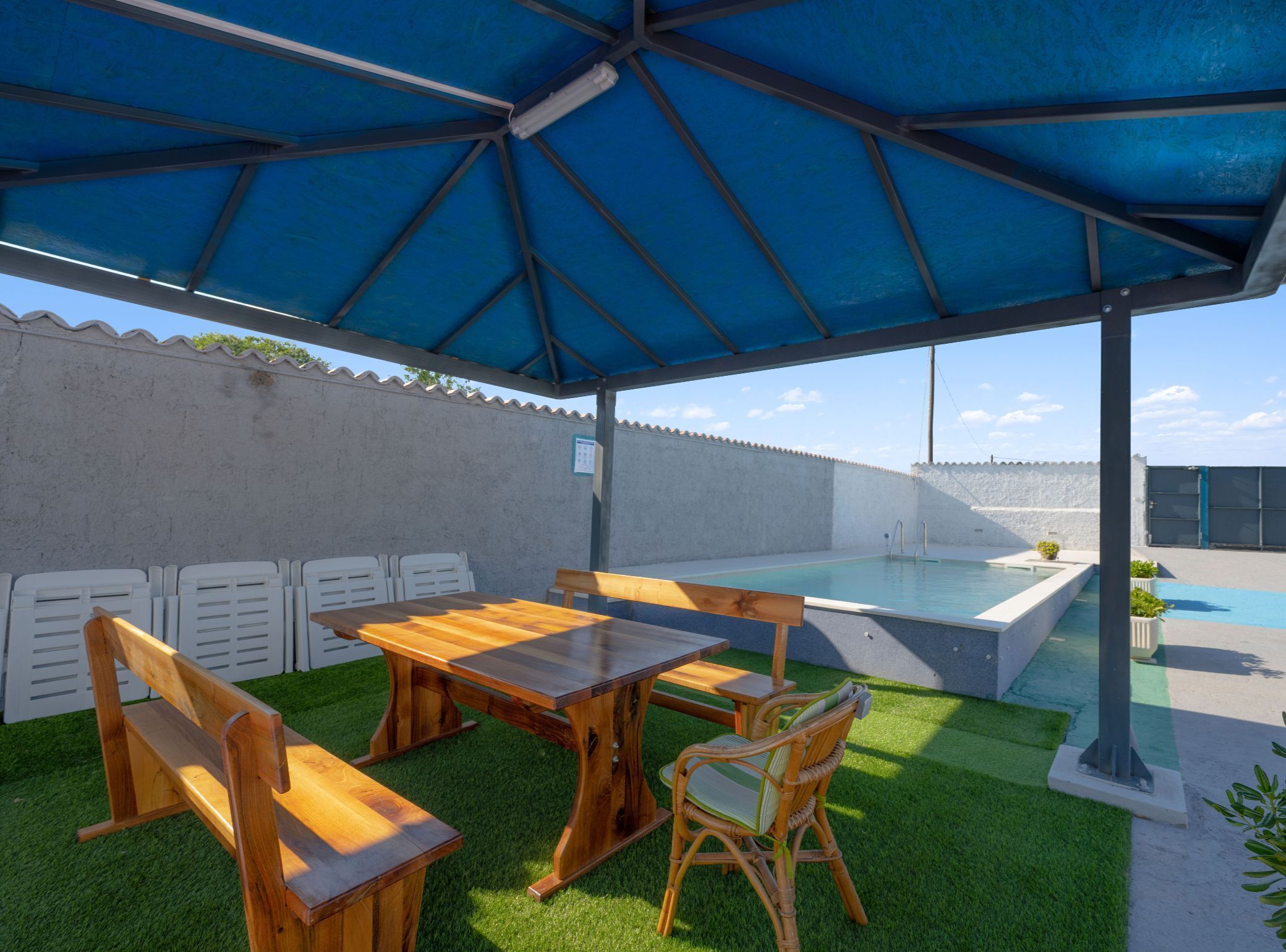 An outdoor patio with a pool, wooden furniture, and a blue canopy.