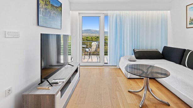 A modern living room with a TV, sofa, glass table, and a balcony with a view of the landscape. - Holiday Home Eros (Sibenik)