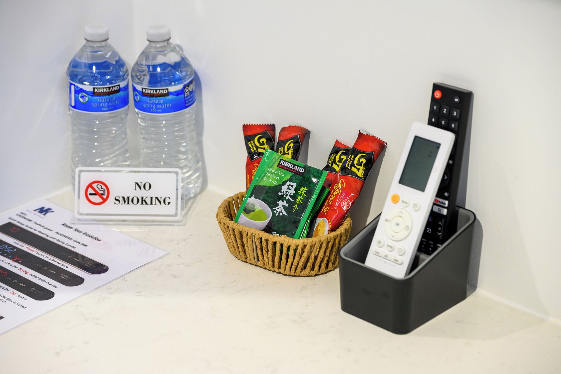 Señal de no fumar, dos botellas de agua mineral Kirkland, una cesta con bolsitas de té verde y café, y una estación de carga para el control remoto.