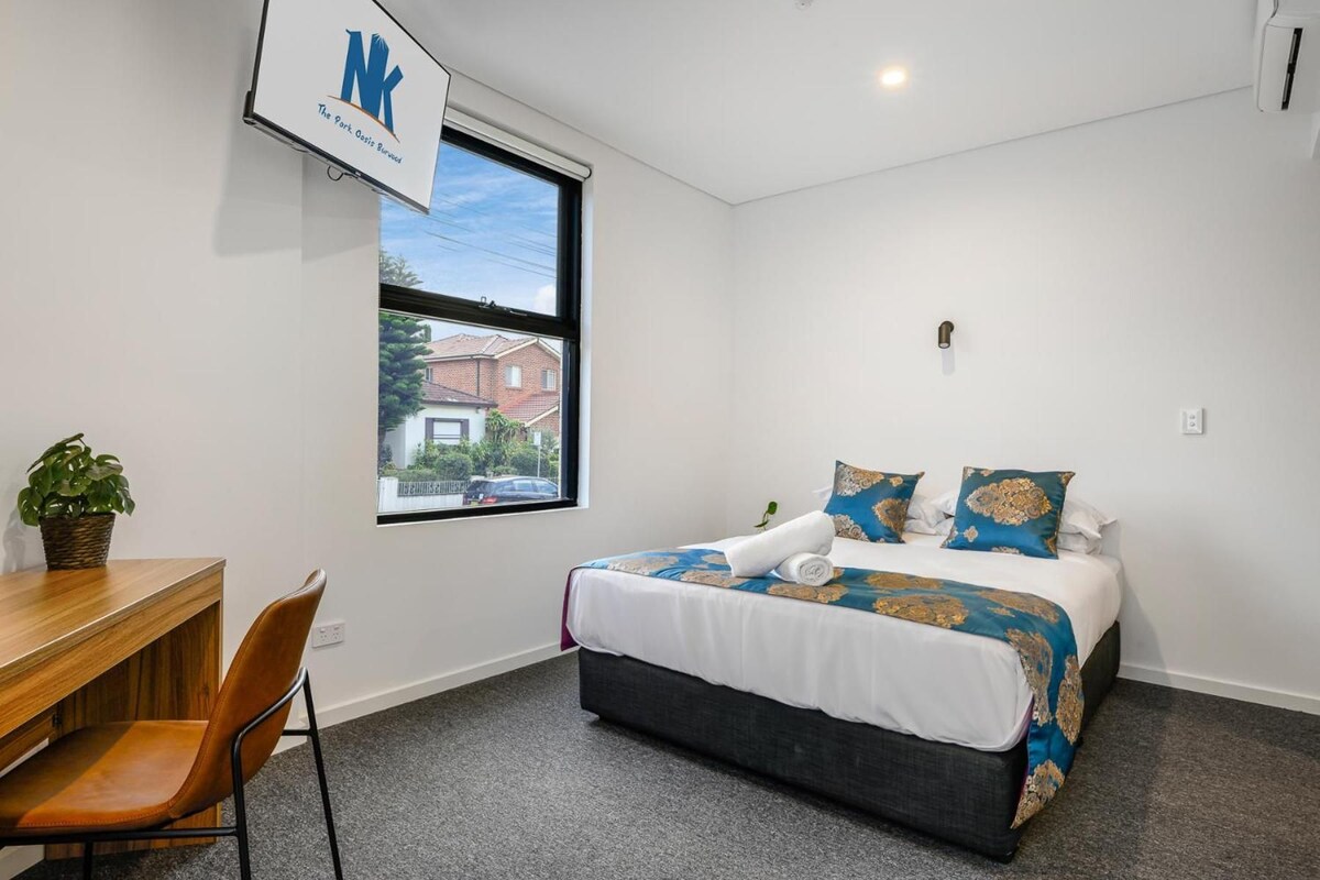 A hotel room with a bed, a desk, a chair, a potted plant, and a window with a view of a house.