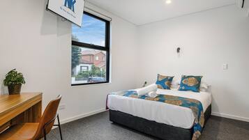 Habitación de hotel con cama, escritorio, silla, planta en maceta y ventana con vista a un edificio.