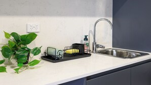 A kitchen with a white countertop, a stainless steel sink, a black dish rack, a green plant, and a power outlet. - THE PARK OASIS BURWOOD (Burwood)