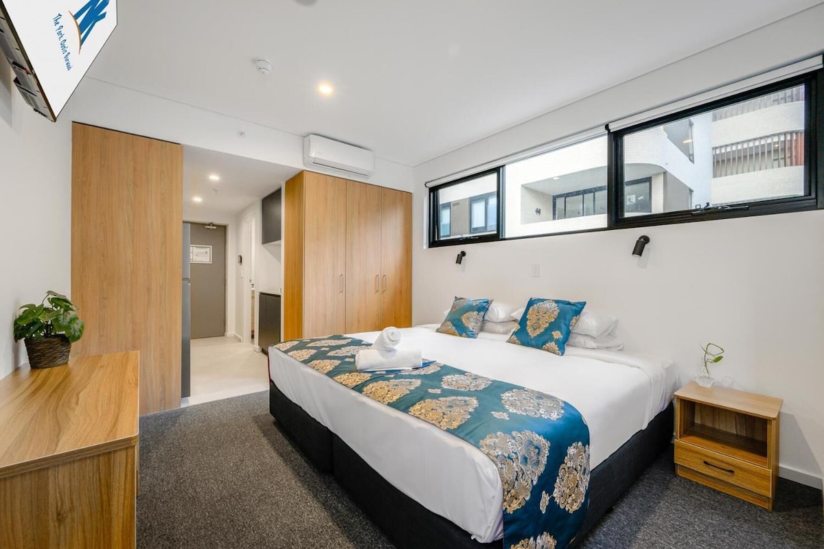 A modern hotel room with a large bed, a small bedside table, a window with a view, and a wooden wardrobe.
