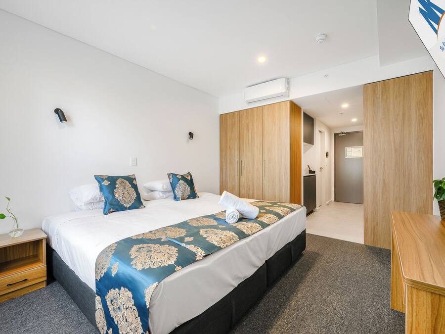 A modern hotel room with a large bed, a wooden wardrobe, a small desk, and a potted plant.