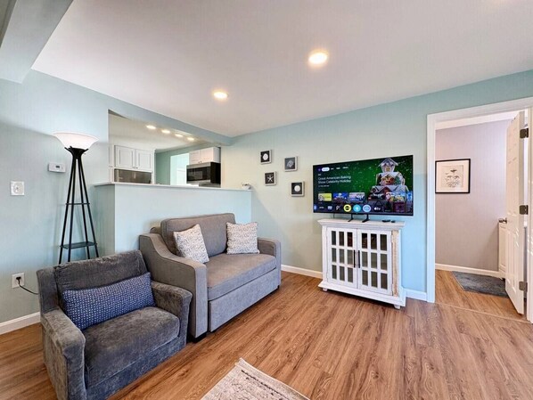 A living room with a grey sofa, a small white cabinet, and a television displaying a show.
