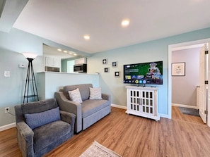 Una sala de estar con un sofá gris, un mueble blanco pequeño y un televisor que muestra un programa.