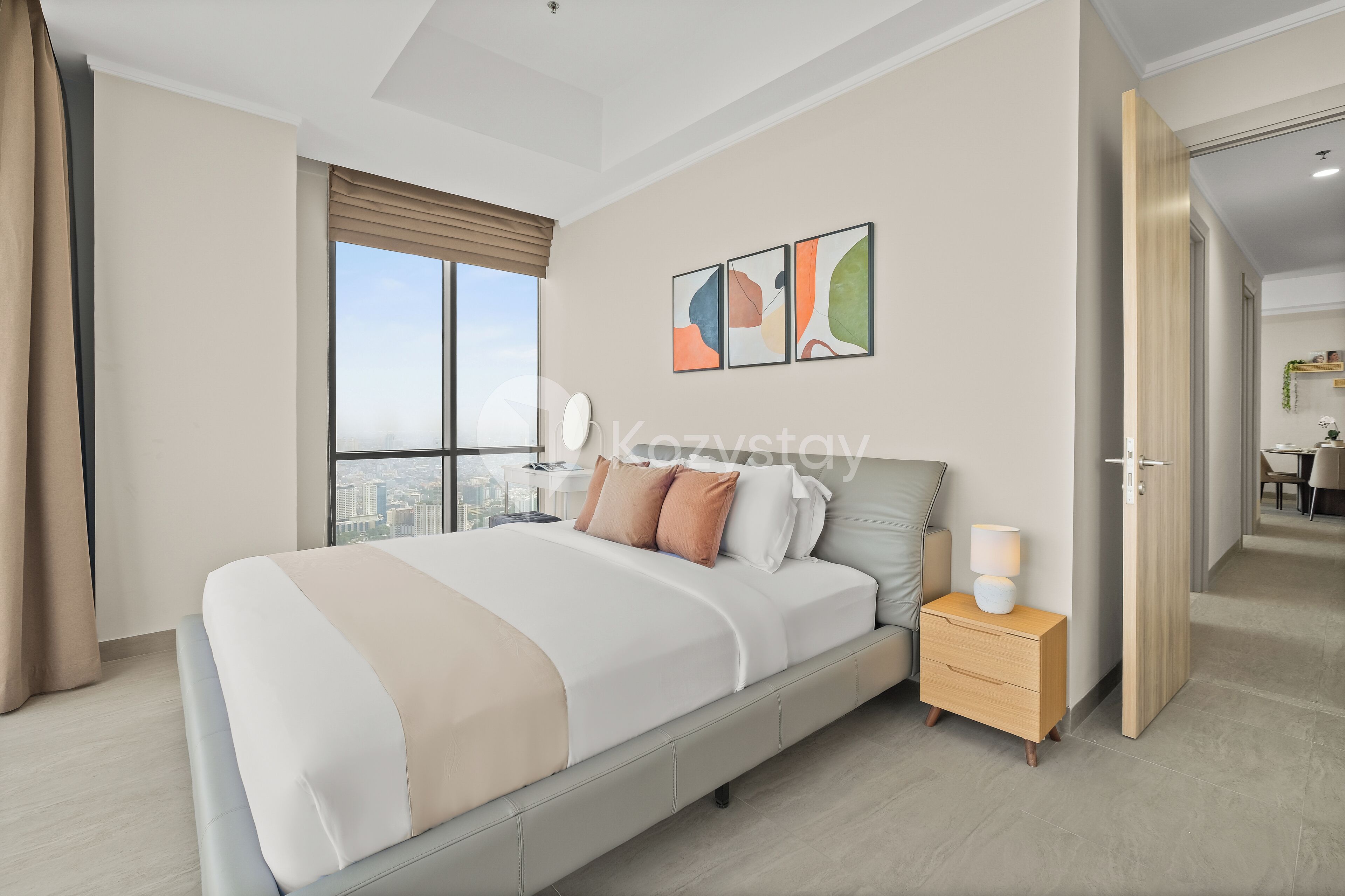 A modern hotel room with a large bed, a nightstand, a lamp, and a city view through the window.