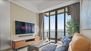 A modern living room with a large flat-screen TV displaying 'NETFLIX', a wooden coffee table, a sofa with patterned cushions, and a large window offering a city view. - Terava by Kozystay - Kemayoran (Jakarta)
