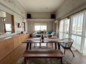 A modern kitchen with wooden cabinets, a dining table with chairs, and a bench. There is a large window with sheer curtains and a view of buildings outside.