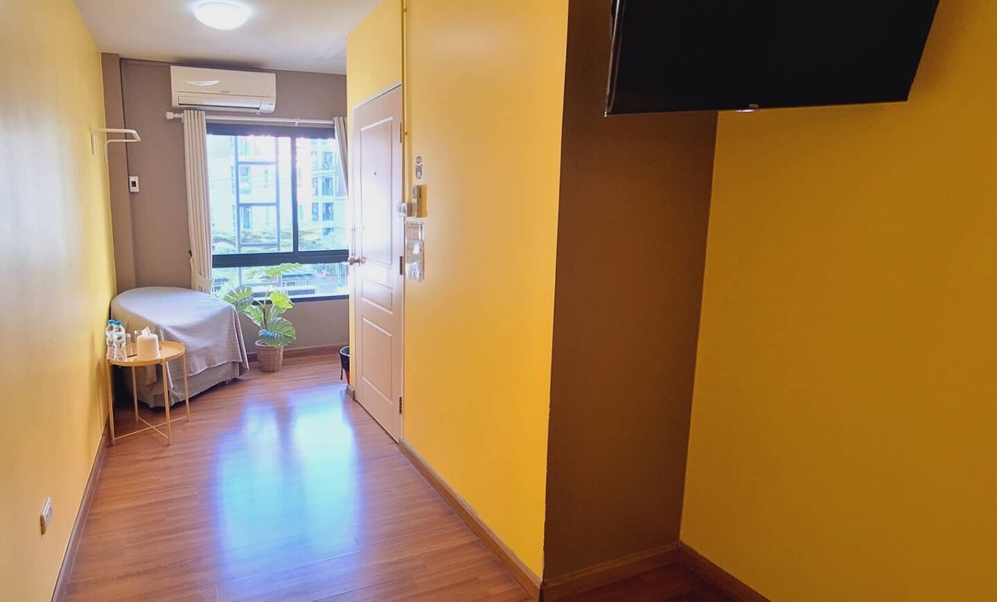 A hallway with wooden flooring, a bed with a white cover, a small table with toiletries, and a window with a view of buildings.