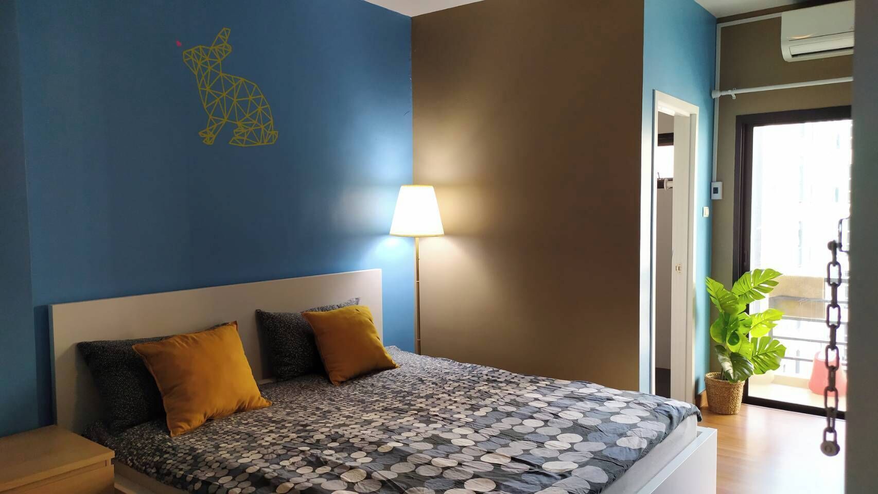 A bedroom with a bed, two pillows, a nightstand, a lamp, and a potted plant.