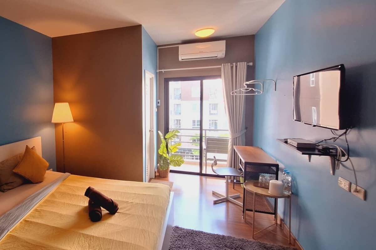 A hotel room with a bed, a desk, a chair, a TV, and a balcony with a view of buildings.