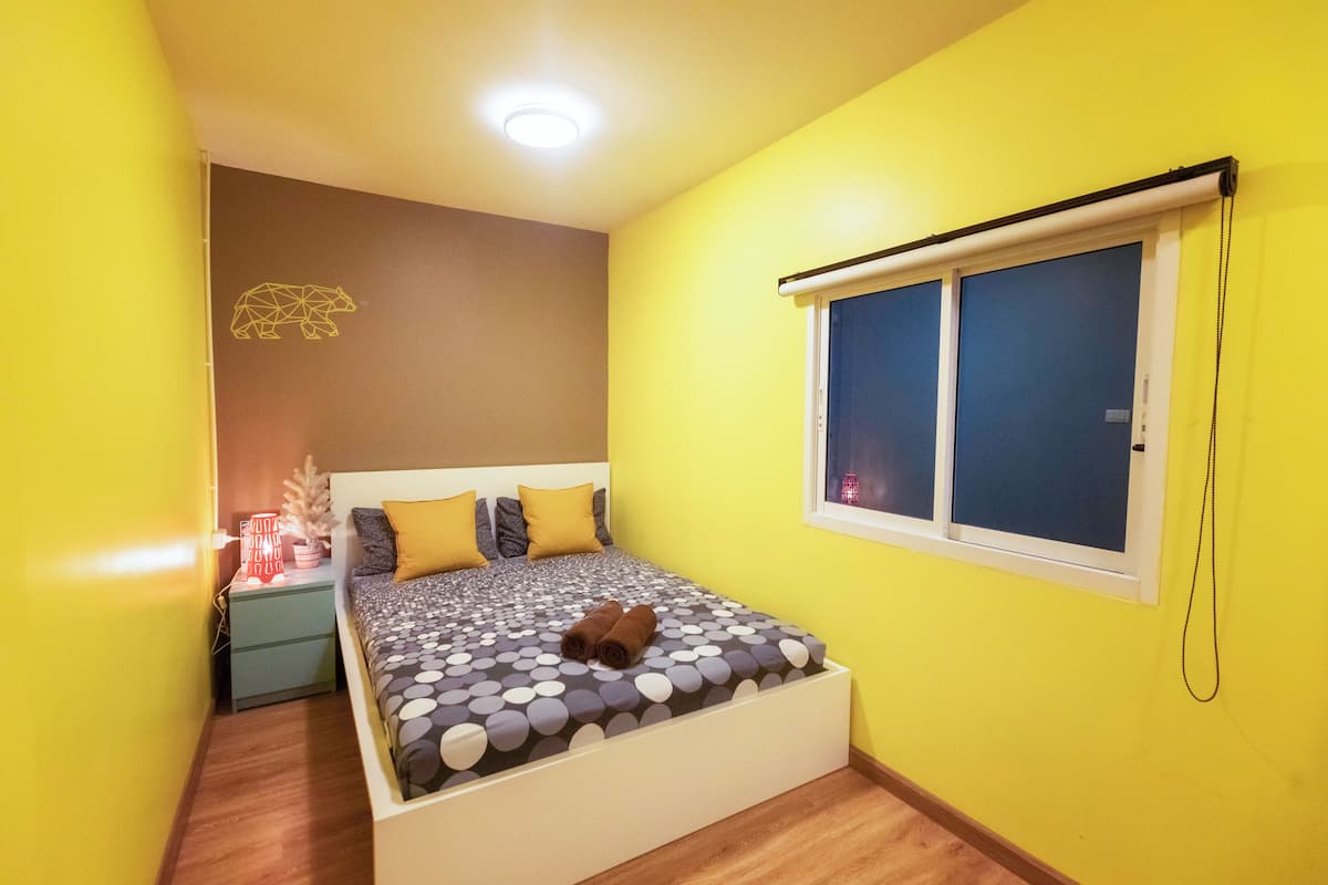 A small, brightly colored bedroom with a bed, a nightstand, a window with blinds, and a wall-mounted light fixture.