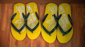 A pair of yellow flip-flops with white leaf patterns and the text "GREEN LINE CH 1002" on a wooden floor. - Peony's Home (Bangkok)