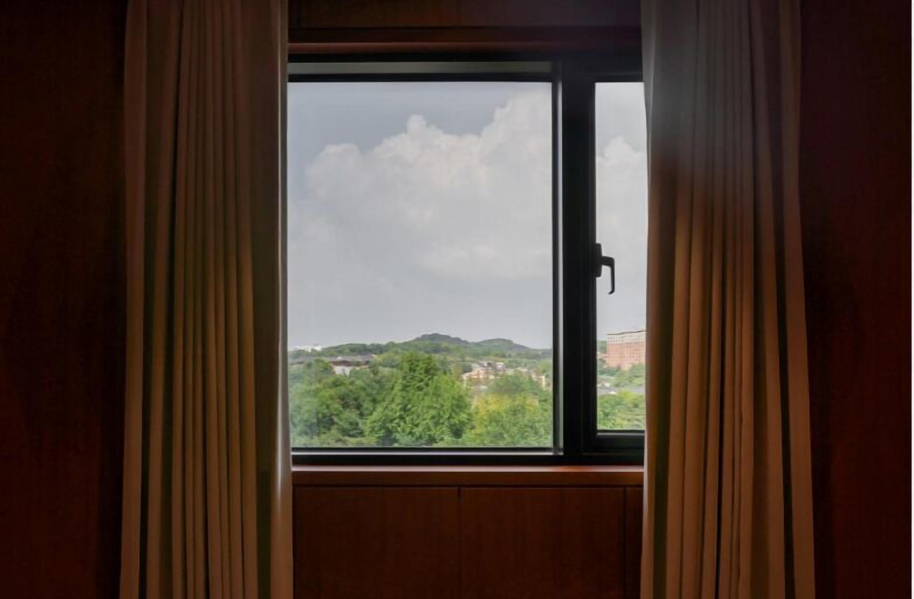 A window with curtains, offering a view of a landscape with trees and hills.