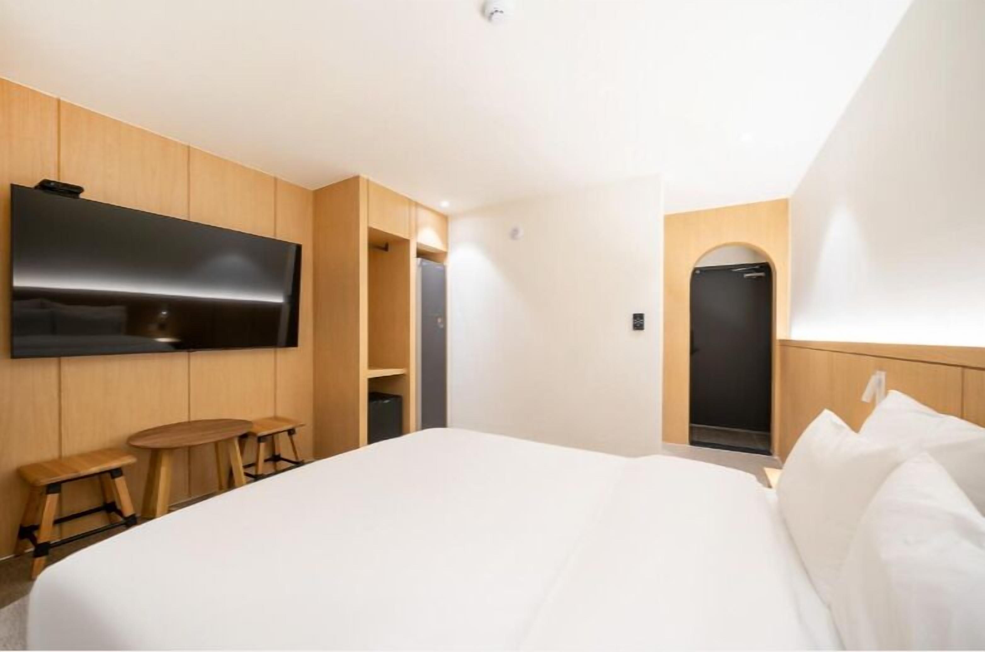 A modern hotel room with a large bed, a flat-screen TV mounted on the wall, wooden furniture, and a built-in wardrobe.