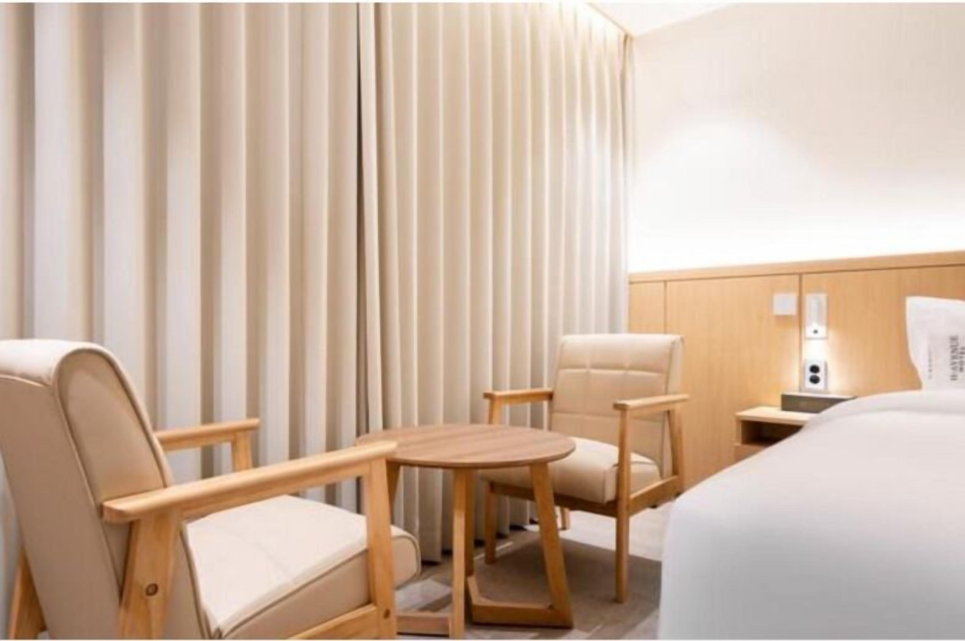 Una habitación de hotel con una cama, dos sillones, una mesita redonda, una mesita de noche con una lámpara y una cortina.