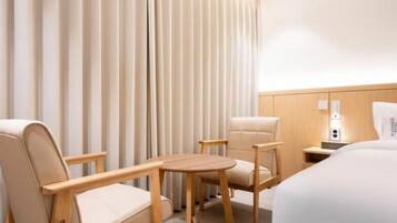 Una habitación de hotel con una cama, dos sillones, una mesita redonda, una mesita de noche con una lámpara y una cortina.