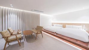 Una habitación de hotel moderna con una cama grande, dos sillones, una mesita y un ventanal amplio con persianas.