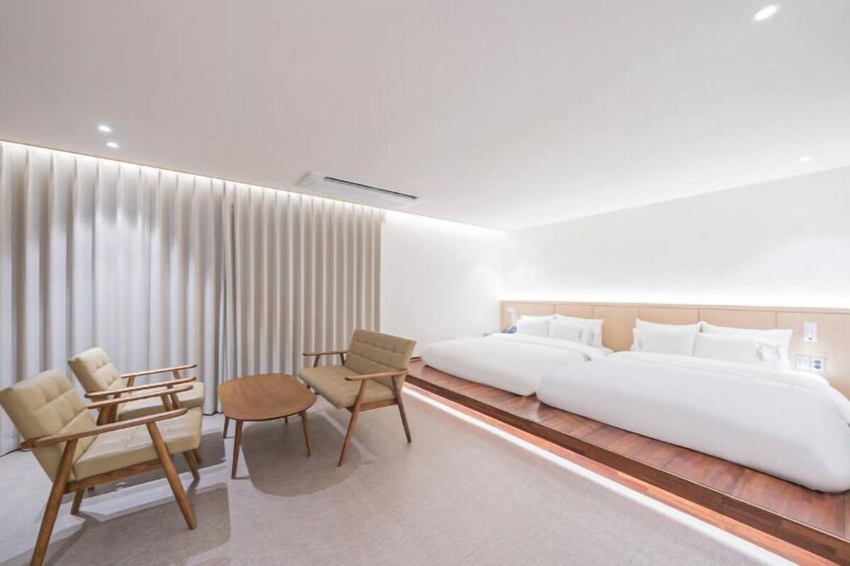 A modern hotel room with a large bed, two armchairs, a small table, and a large window with blinds.