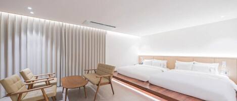 A modern hotel room with a large bed, two armchairs, a small table, and a large window with blinds.