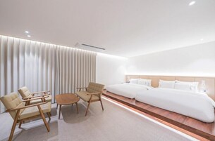 Una habitación de hotel moderna con una cama grande, dos sillones, una mesita y un ventanal amplio con persianas.
