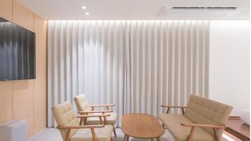 Een moderne woonkamer met een flatscreen televisie aan de muur, twee beige fauteuils, een houten salontafel en een groot raam met verticale jaloezieën.