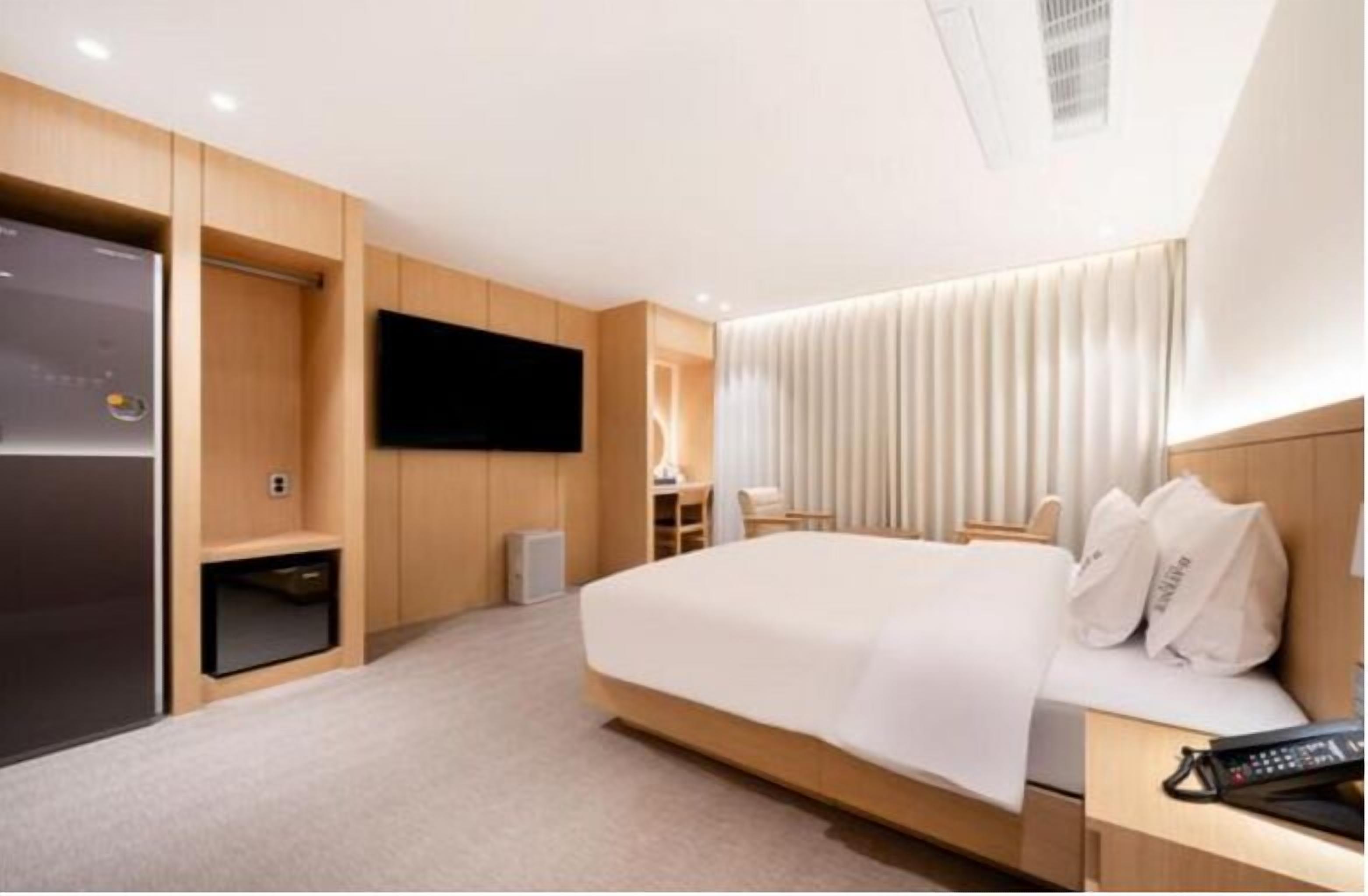 A modern hotel room with a large bed, a flat-screen TV mounted on the wall, a desk with a chair, and a built-in wardrobe.