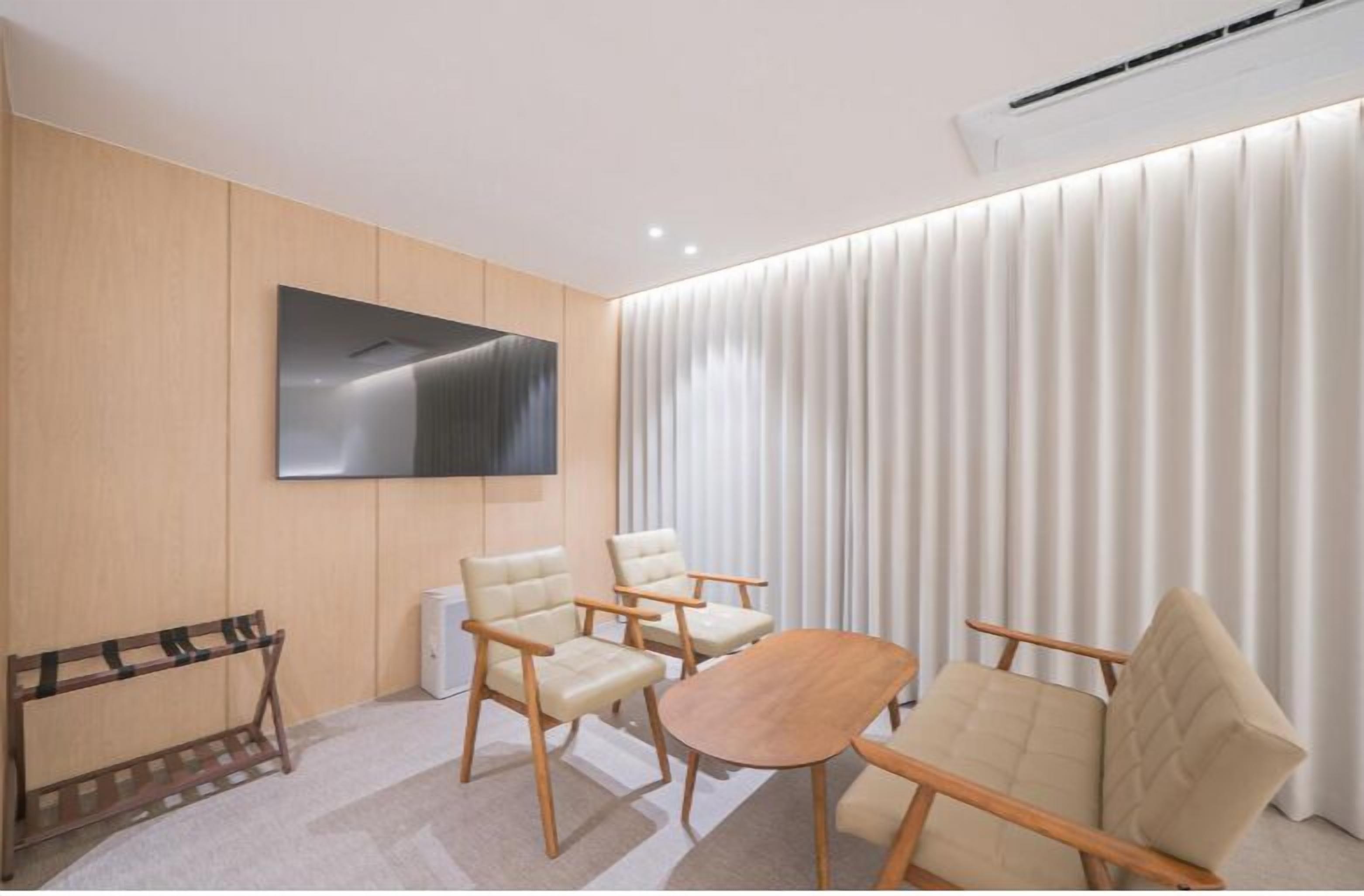 A modern room with a flat-screen TV mounted on the wall, a wooden bench, and three beige armchairs around a small wooden table.