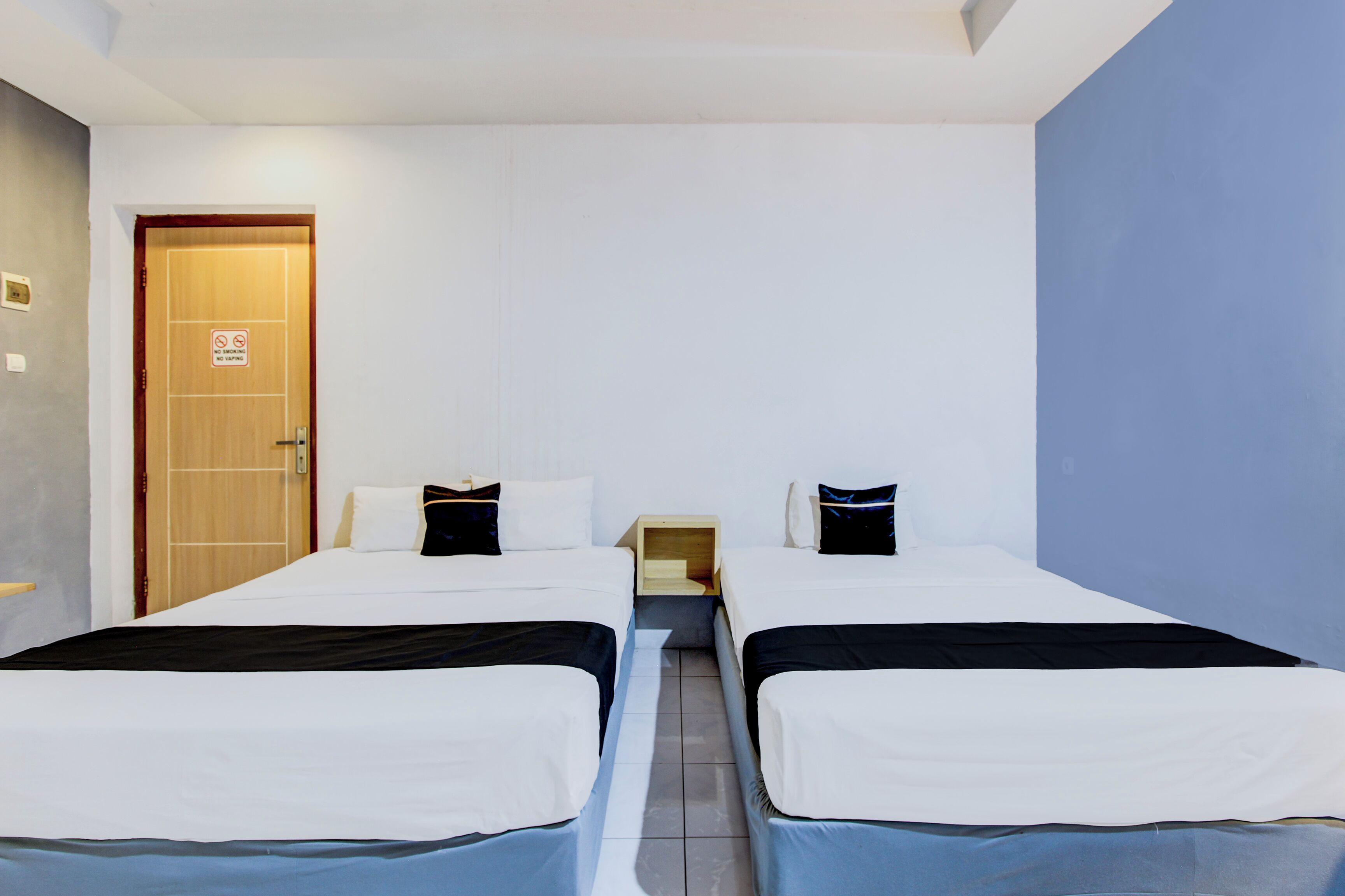 Habitación de hotel con dos camas, una puerta de madera, una mesita de noche pequeña de madera y una pared azul decorativa.