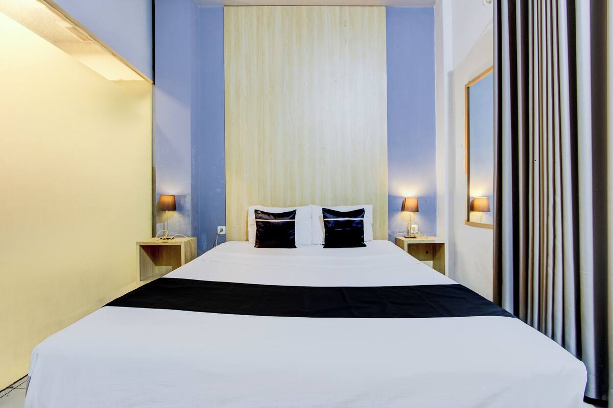 A hotel room with a large bed, two bedside tables with lamps, a window with curtains, and a wall-mounted mirror.