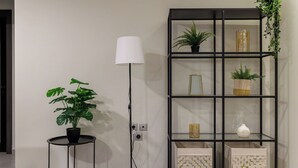 Una habitación moderna con una lámpara de pie negra, una mesa redonda con una planta en maceta y un estante negro con objetos decorativos y cajas de almacenamiento.