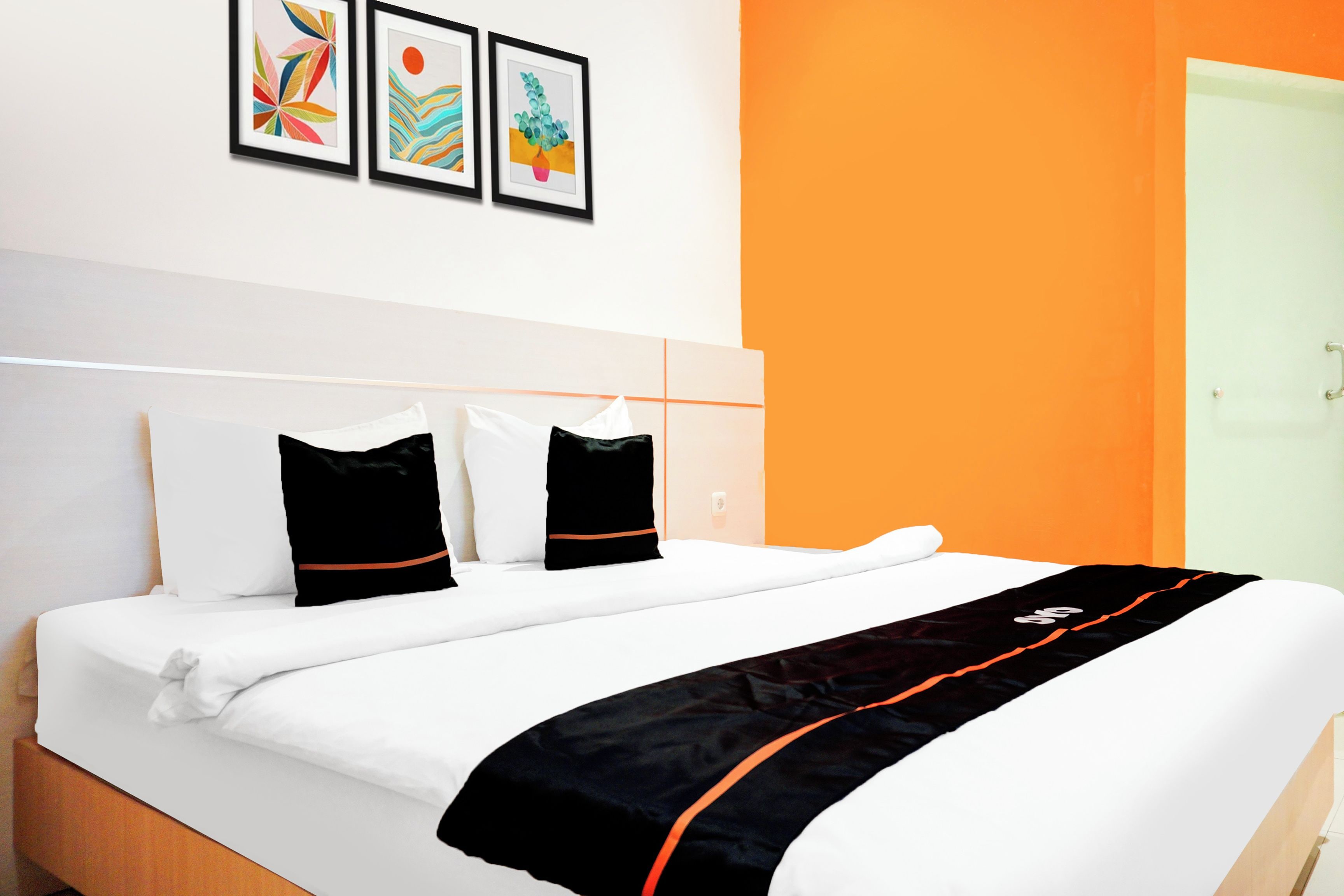 A hotel room with a bed, white bedding, two black pillows, and a black and orange throw. There are three framed artworks on the wall.