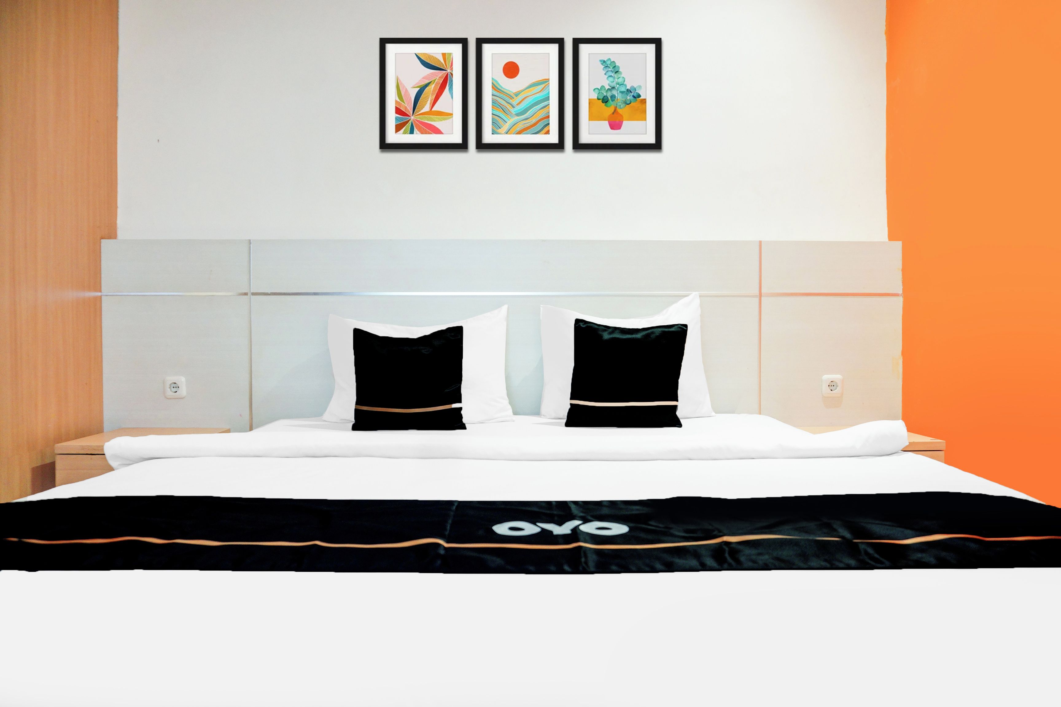 A hotel room with a bed, two pillows, a headboard, and three framed artworks on the wall.