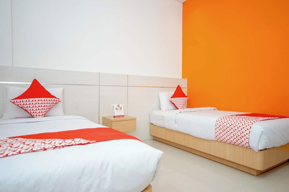 Quarto de hotel com duas camas, cada uma com roupa de cama branca e almofadas vermelhas. Há uma pequena mesa de cabeceira com uma luminária e um livro. O quarto tem uma paleta de cores branca e laranja.