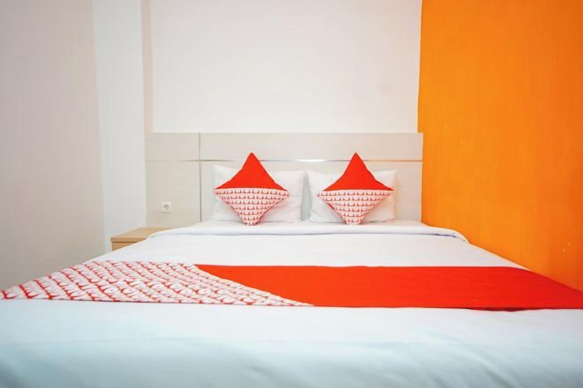 Ein Bett mit weißer Bettwäsche, zwei roten Kissen und einer roten Tagesdecke, sowie eine weiße Wand mit orangefarbenem Akzent.