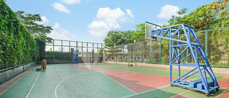 An outdoor basketball court with a blue metal structure, surrounded by greenery and a clear sky.