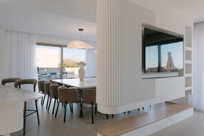 Una sala de estar moderna con un televisor de pantalla plana grande montado en una pared blanca, una zona de comedor con una mesa y sillas de mármol, y vistas a la ciudad a través de amplias ventanas.