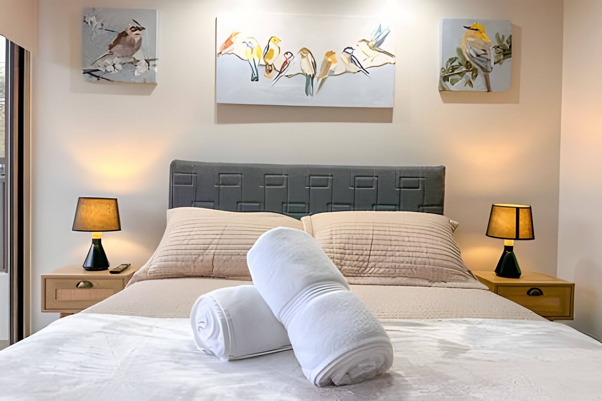 Habitación de hotel con cama, dos lámparas de noche, un cuadro grande de pájaros en la pared y una ventana a la izquierda.