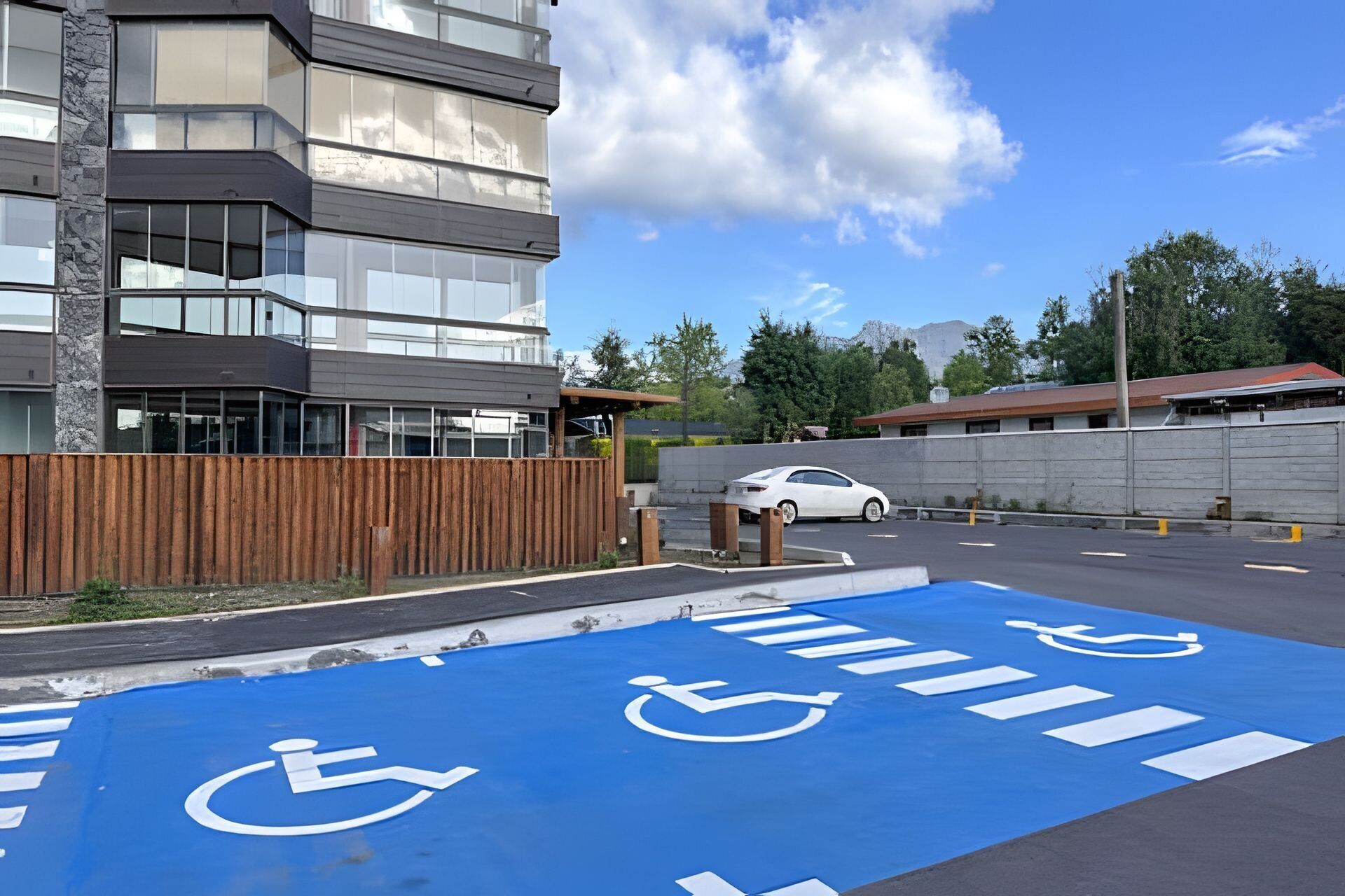 Un área de estacionamiento con espacios reservados para discapacitados, identificados con pintura azul, y un coche blanco estacionado cerca.