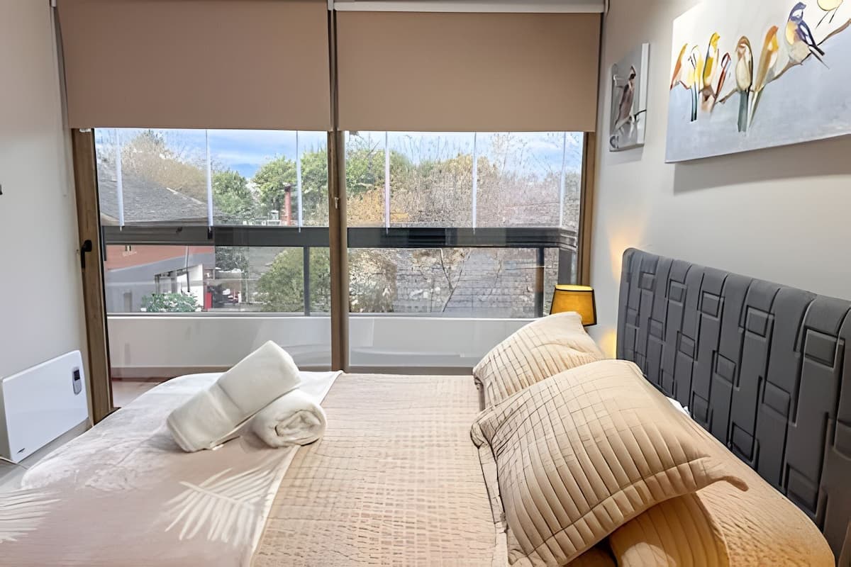 A bedroom with a large window, a bed with beige bedding, and a grey headboard.