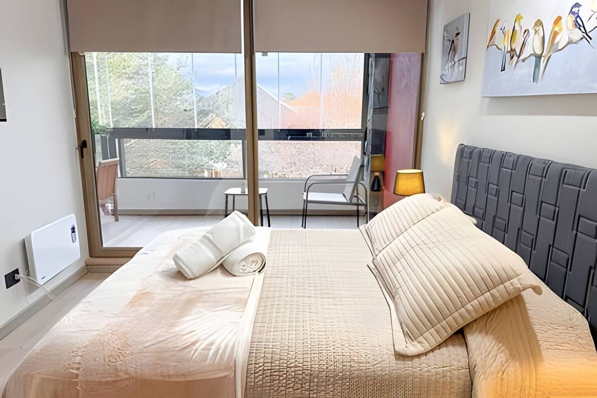 A hotel room with a bed, a window with a view of the outdoors, a lamp, and artwork on the wall.