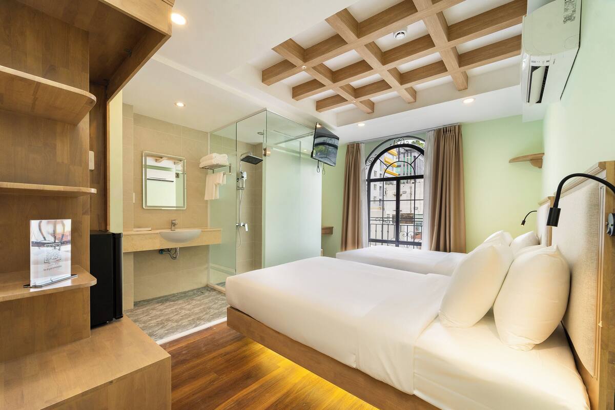 A modern hotel room with a wooden ceiling, a large bed with white bedding, a glass shower enclosure, and a view of the outside through a window with curtains.