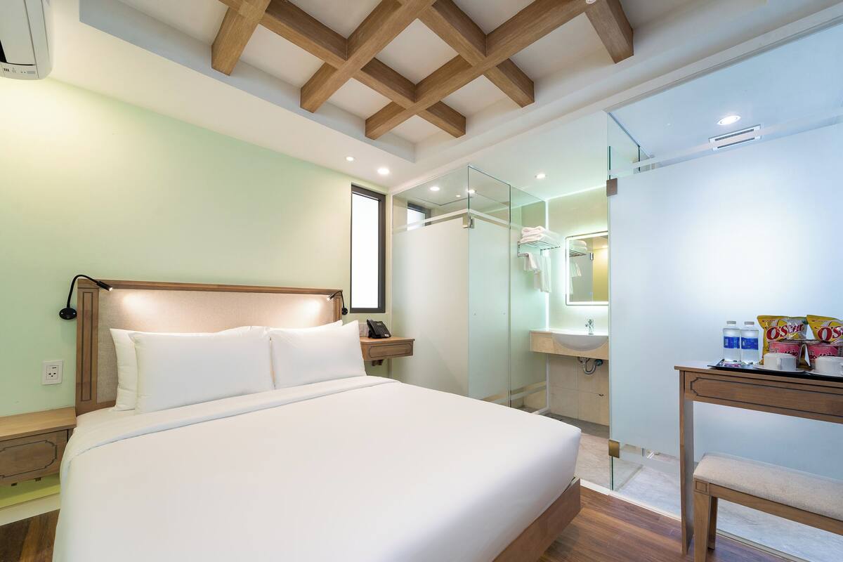 A hotel room with a large bed, a wooden headboard, a bedside table with a lamp, a mirror, a small table with toiletries, and a bathroom visible through a glass partition.
