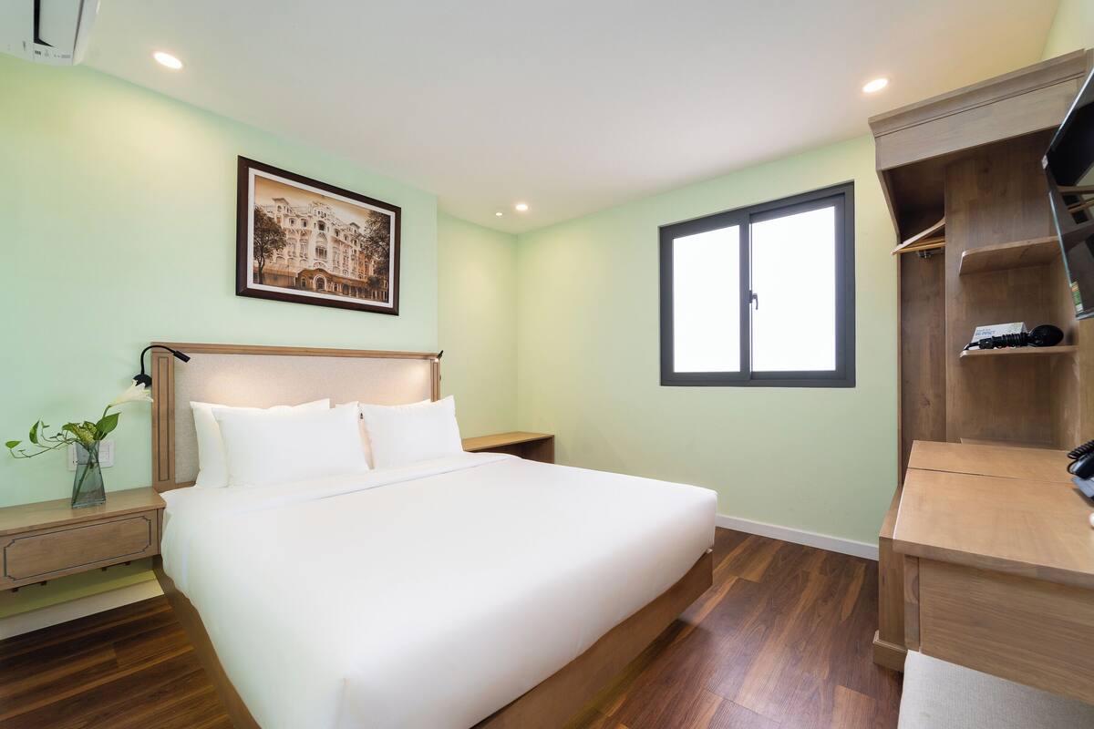 A bedroom with a bed, a nightstand, a wooden wardrobe, a window, and a framed picture on the wall.