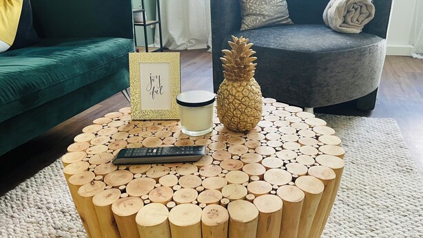 Une table basse ronde en bois, sur laquelle se trouvent une télécommande et une bougie, entourée d’un tapis tissé.