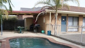 A swimming pool with a brick border, a covered patio area with green chairs, and a fenced backyard. - Country Home Motor Inn (Shepparton)