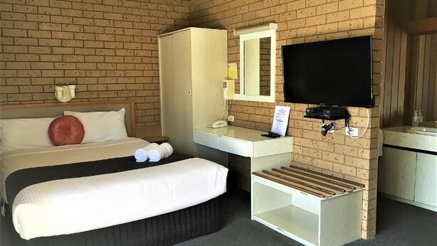 A hotel room with a brick wall, a bed with white bedding, a desk with a mirror, a television, and a small table. - Country Home Motor Inn (Shepparton)